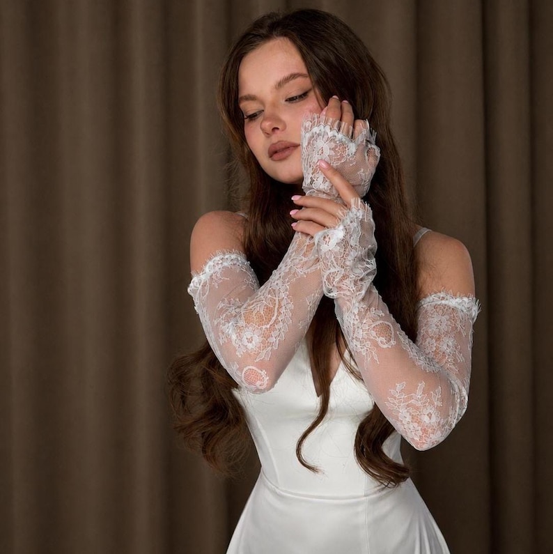 Puede incluir: Guantes de novia de encaje blanco con un delicado dise&ntilde;o floral. Los guantes son largos, se extienden m&aacute;s all&aacute; de los codos y tienen un borde festoneado. La tela transparente permite que se vea la piel, creando un aspecto elegante.