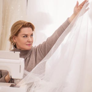 Puede incluir: Una mujer con un jersey de cuello alto beige trabaja con tela blanca cerca de una m&aacute;quina de coser. Sostiene una tela blanca transparente y brillante, probablemente tul, con su mano derecha. La escena sugiere costura o confecci&oacute;n.