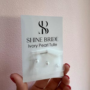 Puede incluir: Una tarjeta blanca con el texto "SHINE BRIDE Ivory Pearl Tulle" presenta un logotipo y tres pequeñas perlas redondas de color marfil unidas a un trozo de tul blanco. La tarjeta se sostiene sobre un fondo rosa.