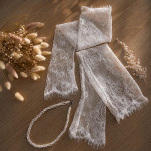 Puede incluir: Una composici&oacute;n de accesorios nupciales sobre una superficie de madera clara. Incluye una bufanda de encaje blanco, un collar de perlas y un adorno para el cabello de perlas, con flores secas.