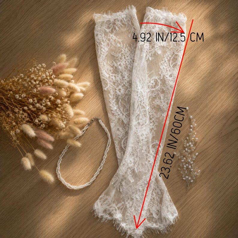 Puede incluir: Un par de mangas de encaje blanco, de 60 cm de largo, con un delicado estampado floral y bordes festoneados. Las mangas se exhiben sobre una superficie de madera con un collar de perlas y flores secas.