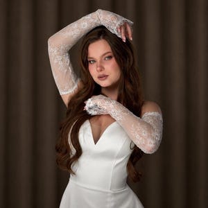 Puede incluir: Un vestido de novia de sat&eacute;n blanco con escote en V. El vestido se combina con guantes largos de encaje transparente. Los guantes presentan un delicado estampado floral y se extienden hasta la parte superior de los brazos. La modelo tiene el pelo largo y casta&ntilde;o.