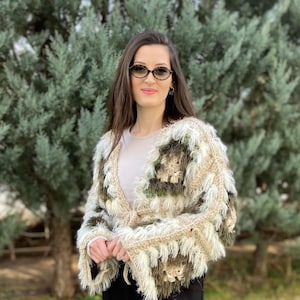 May include: A handmade, open-front crochet jacket with a shaggy, fringed texture. The jacket features a color block design in shades of beige, olive green, and cream. The sleeves are flared, and the jacket is tied at the waist.
