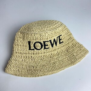 May include: A beige woven bucket hat with the word "LOEWE" embroidered in black across the front. The hat has a wide brim and a slightly rounded crown. The hat is made of a natural fiber material.