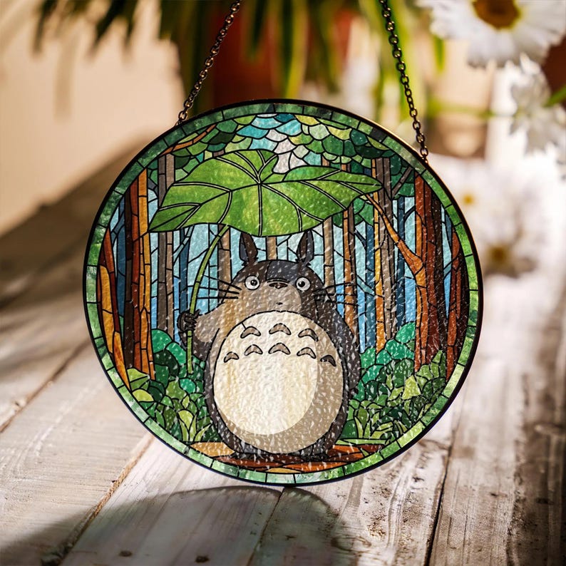 May include: Round stained glass window featuring a cartoon character under a large leaf in a forest scene. The window has a black metal frame and a chain for hanging. The design incorporates shades of green, blue, brown, and white.