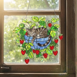 May include: A stained glass window decoration featuring a tabby cat sleeping in a blue basket surrounded by red strawberries and green leaves. The window art is suspended by a silver chain, creating a charming, whimsical scene.