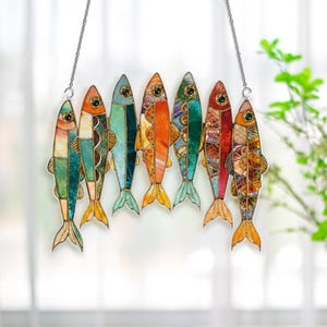 May include: A decorative hanging featuring seven colorful fish. Each fish has a unique stained-glass design with shades of green, blue, orange, and red. The fish are suspended by a silver chain, creating a vibrant and artistic display.
