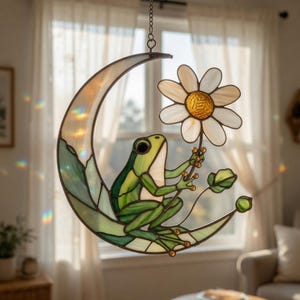 May include: Stained glass suncatcher featuring a green frog holding a daisy, perched on a crescent moon. The moon has a gradient of colors, and the daisy has a yellow center. The suncatcher hangs from a chain.