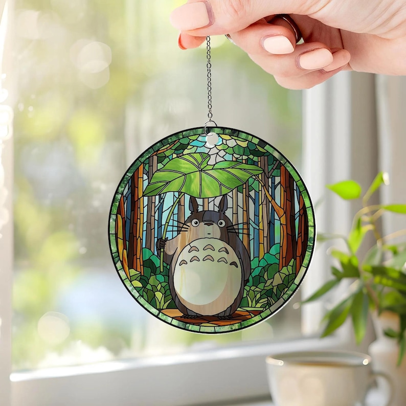May include: A round stained glass ornament featuring a cartoon character holding a leaf umbrella in a forest scene. The ornament has a chain for hanging and is held up by a hand.