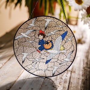 May include: Round stained glass window featuring a young girl on a broom with a black cat, surrounded by seagulls. The girl wears a blue dress and a red bow. The window has a textured, light-colored background and a black metal frame.