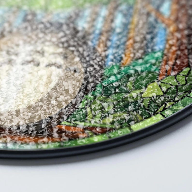 May include: Round decorative coaster with a mosaic design featuring a forest scene. The coaster has a black border and a textured surface. The artwork includes shades of green, brown, gray, and blue.