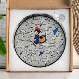 May include: Round stained glass window featuring a girl on a broom with a black cat, surrounded by seagulls. The girl wears a blue dress and a red bow. The window has a black metal frame and a suction cup for hanging.