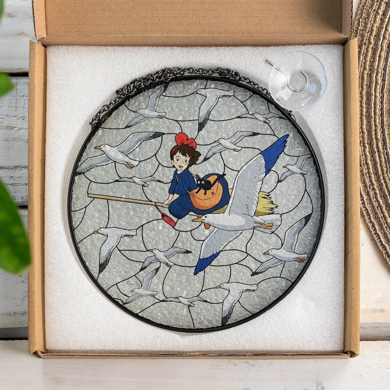 May include: Round stained glass window featuring a girl on a broom with a black cat, surrounded by seagulls. The girl wears a blue dress and a red bow. The window has a black metal frame and a suction cup for hanging.