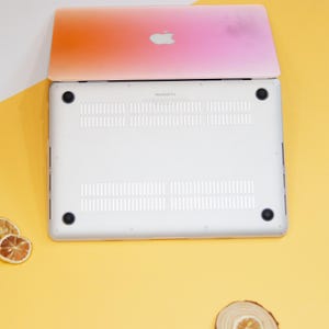 Puede incluir: La parte inferior de un port&aacute;til plateado con un dise&ntilde;o degradado rosa y naranja en la tapa. El port&aacute;til est&aacute; sobre una superficie amarilla.