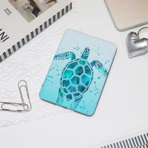 May include: A light blue tablet case featuring a teal and turquoise sea turtle design. The turtle is surrounded by splashes of water. The case is rectangular and has rounded corners. The background is a gradient of light blue.