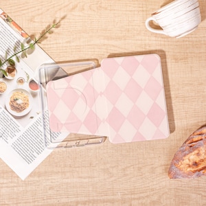 May include: A clear plastic protective case for an e-reader or tablet featuring a light pink and white diamond argyle pattern on the front cover. The case is rectangular with rounded corners and a circular cutout on the interior left side panel.