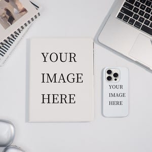 May include: A white notebook and phone case with the text "YOUR IMAGE HERE" are displayed on a white surface, alongside a laptop and a book. The notebook and phone case are blank, ready for customization.