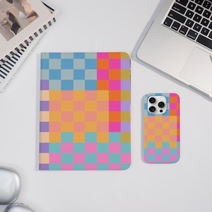 May include: A notebook and phone case set with a colorful checkerboard pattern in shades of blue, pink, orange, and purple. The notebook is open, and the phone case is next to it. A laptop is in the background.