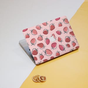Op de afbeelding: Een laptop met een roze hoes met een patroon van aardbeien in verschillende tinten rood en roze, omlijnd in zwart. De laptop staat op een wit en geel oppervlak.