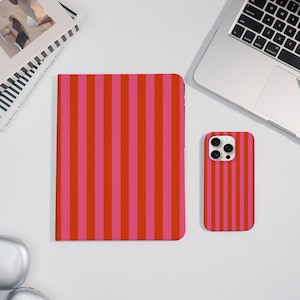 Op de afbeelding: Een rood-roze gestreepte telefoonhoes en een bijpassend notitieboek worden gepresenteerd op een wit oppervlak. De telefoonhoes staat naast een laptop en een gesloten notitieboek. Het notitieboek en de telefoonhoes hebben een verticaal streepjespatroon.