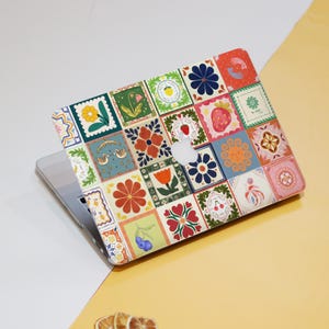 May include: A laptop with a colorful, patterned cover. The cover features a patchwork design of squares with floral, fruit, and bird illustrations in various colors. The laptop is on a white and yellow surface.