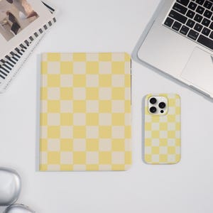 May include: A yellow and cream chequered pattern notebook and phone case are displayed on a white surface. The notebook is closed and rectangular. The phone case is a rectangle with rounded corners. A laptop is partially visible.