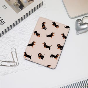 Peut inclure: Une housse de tablette rose clair avec un motif répété d'illustrations de teckels noirs et fauves. Les teckels sont dans diverses poses. L'étui est rectangulaire et semble être fait d'un matériau lisse. D'autres articles sur la surface blanche incluent un livre et un cœur argenté.