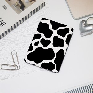 May include: A tablet case with a black and white cow print design. The case has a rectangular shape with rounded corners and a repeating pattern of black blotches on a white background. Other items on the white surface include a silver heart and a book.