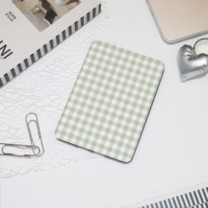May include: A rectangular, light green and white gingham patterned notebook or notepad. The notebook is on a white surface, with a silver heart-shaped object and a silver paperclip nearby. A book with black and white stripes is also visible.