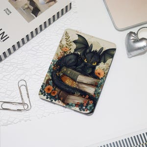 May include: A rectangular phone case featuring a black dragon illustration. The dragon is lying on a stack of books, surrounded by flowers and foliage. The case has a cream-colored background and a glossy finish.