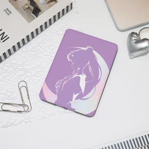Peut inclure: Une coque de tablette lavande avec une silhouette blanche d'une femme aux longs cheveux et un motif de croissant de lune. L'étui est rectangulaire avec des coins arrondis. Un objet argenté en forme de cœur et un livre sont également visibles.