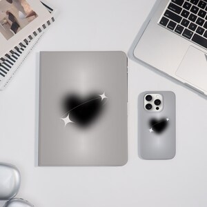 May include: A gray tablet case and phone case with a black heart design and white star accents. The cases are on a white surface next to a laptop and a book.