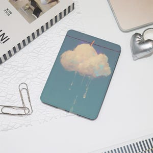 May include: A teal phone case featuring a cloud illustration with rain drops. The cloud is light yellow and white, with a small wooden clothespin attached to a purple line. The case is on a white surface with a silver heart-shaped object.