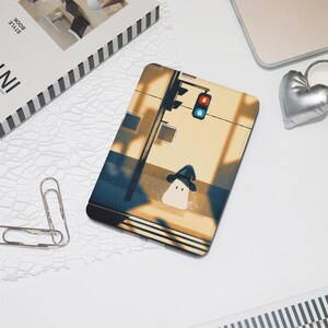 May include: A rectangular phone case featuring an illustration of a street scene with a traffic light and a cartoon character wearing a hat. The case has a beige and blue color scheme with a black border. Other items include a silver heart-shaped object and a book.