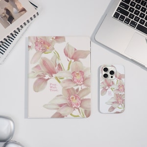May include: A notebook and phone case with a pink orchid floral design. The notebook and phone case have the words "Pray. Wait. Trust." printed on them. A laptop and a book are also in the image.