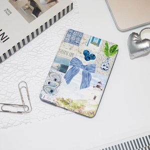 Könnte beinhalten: Ein dekoratives Notizbuch mit einem Collage-Design mit Küstenelementen. Das Design umfasst eine blau gestreifte Schleife, Blaubeeren, einen Leuchtturm und den Text "COASTAL LIFE". Das Notizbuch ist von einem weißen Hintergrund umgeben.