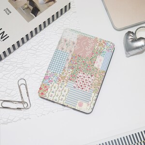 May include: A rectangular notebook with a patchwork design featuring floral, gingham, and striped patterns in pastel colors. The notebook is surrounded by a white surface, a silver heart-shaped object, and a book.