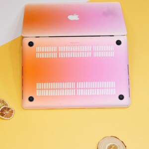 Puede incluir: Un port&aacute;til rosa y naranja con un logotipo de Apple blanco en la tapa. El port&aacute;til est&aacute; cerrado y se ve la parte inferior del port&aacute;til, que muestra los orificios de ventilaci&oacute;n y dos pies de goma negros.