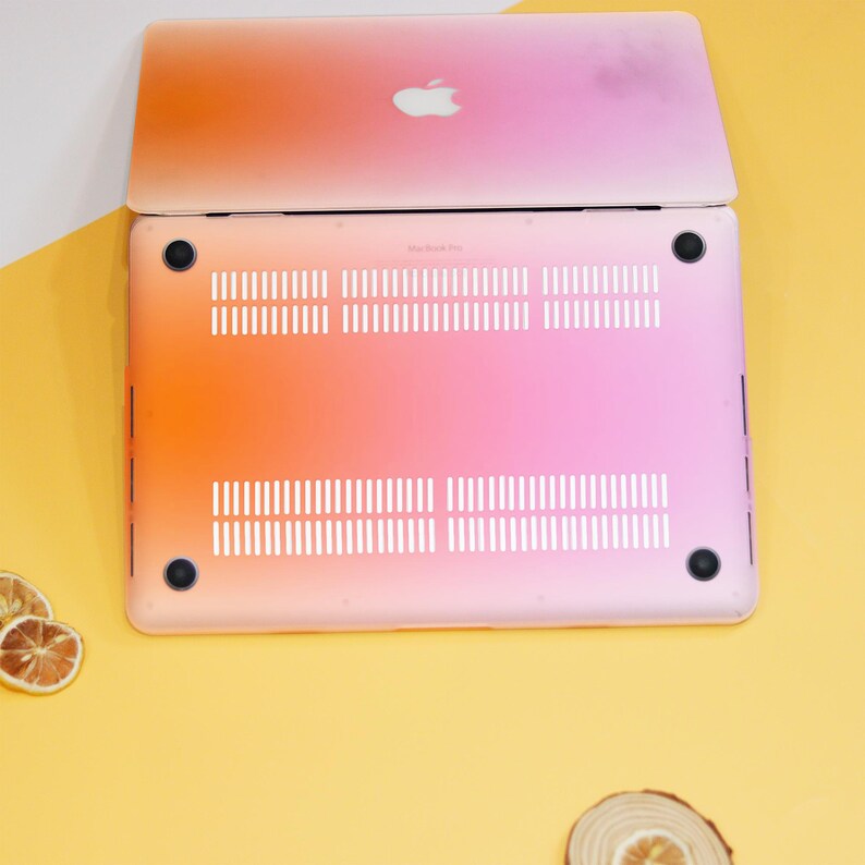 Puede incluir: Un port&aacute;til rosa y naranja con un logotipo de Apple blanco en la tapa. El port&aacute;til est&aacute; cerrado y se ve la parte inferior del port&aacute;til, que muestra los orificios de ventilaci&oacute;n y dos pies de goma negros.
