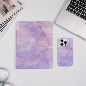 Può includere: Un set di quaderno e custodia per telefono con un design ad acquerello nei toni del viola e della lavanda. Il quaderno è rettangolare e la custodia del telefono ha un ritaglio per la fotocamera. Gli articoli sono esposti su una superficie bianca.