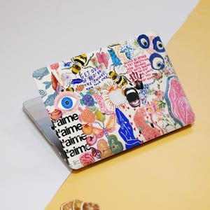Könnte beinhalten: Ein Laptop mit einer farbenfrohen, gemusterten Hülle. Das Design umfasst Bienen, Blumen, Augen und Text, der lautet: "Get out of bed! Do the things! Own your life!" und "Never be afraid to try something new".