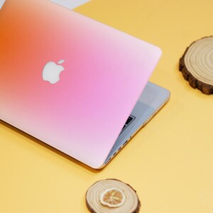 Puede incluir: Una piel para port&aacute;til con degradado rosa y naranja con el logotipo de Apple en la tapa. El port&aacute;til est&aacute; cerrado y sobre una superficie amarilla.