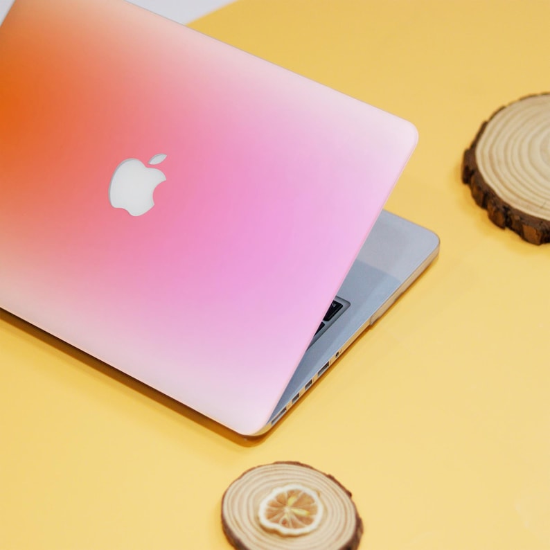 Puede incluir: Una piel para port&aacute;til con degradado rosa y naranja con el logotipo de Apple en la tapa. El port&aacute;til est&aacute; cerrado y sobre una superficie amarilla.