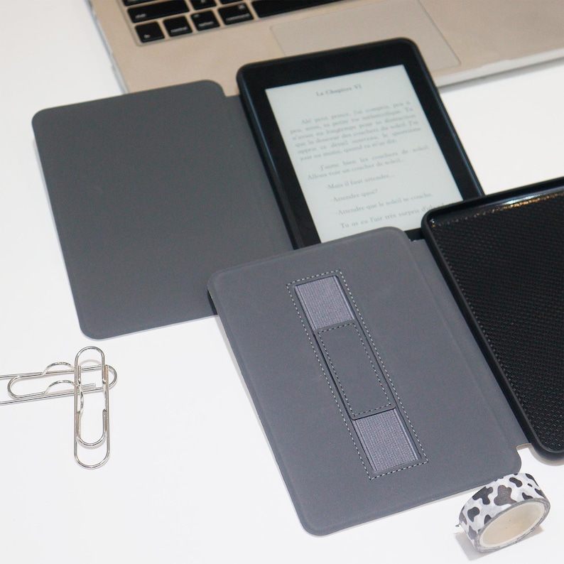 K&ouml;nnte beinhalten: Grauer E-Reader mit schwarzem Bildschirm, der Text anzeigt, ge&ouml;ffnet in einer grauen H&uuml;lle. Die H&uuml;lle hat einen eingebauten St&auml;nder und befindet sich neben einem Laptop, B&uuml;roklammern und einer Rolle dekorativen Klebebands.
