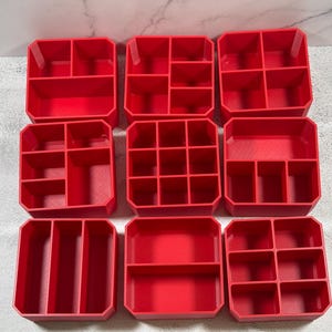 May include: A collection of nine red plastic storage organizers arranged in a three-by-three grid. Each container features a distinct internal compartment layout, including various square, rectangular, and grid-style dividers for sorting small items.