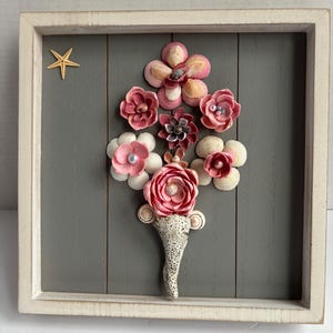 May include: A framed seashell art piece with a gray background. The seashells are arranged to form pink flowers with a starfish in the top left corner.