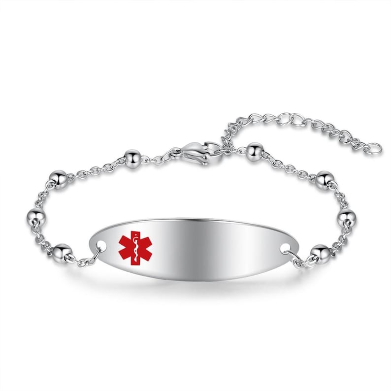 May include: A silver-toned medical alert bracelet with a polished finish. The oval-shaped plate features a red medical symbol. The bracelet has a chain with small, round beads and an adjustable clasp.