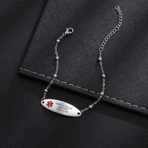 May include: A silver-colored medical ID bracelet with a white oval tag. The tag has a red medical symbol and the words "DIABETES INSULIN WARFARIN PENICILLINP" in black. The bracelet is on a black surface.