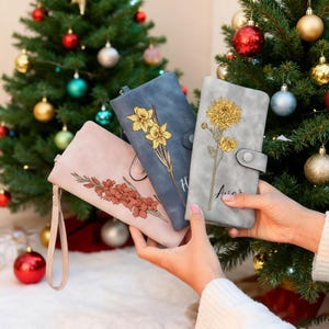 Op de afbeelding: Drie portemonnees met bloemenprint in roze, blauw en grijs worden voor een kerstboom gehouden. De portemonnees hebben een drukknoopsluiting en een polsbandje. De roze portemonnee heeft een bruin bloemmotief, de blauwe gele narcissen en de grijze een gele chrysant.