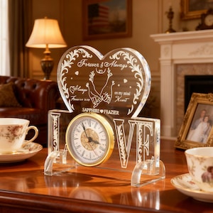 May include: A clear acrylic clock with a gold-tone clock face and the word "LOVE" etched into the base. The clock features a heart-shaped top with the words "Forever & Always" and a couple holding hands. The clock is on a wooden surface.